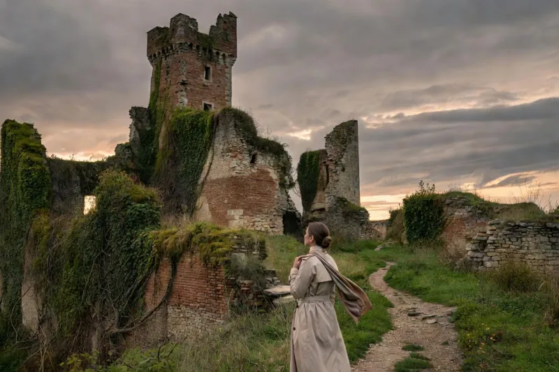 Les murs de brique et de pierre d'un ancien château envahis par le lierre, sous un ciel de soirée, avec une visiteuse au loin sur un chemin.